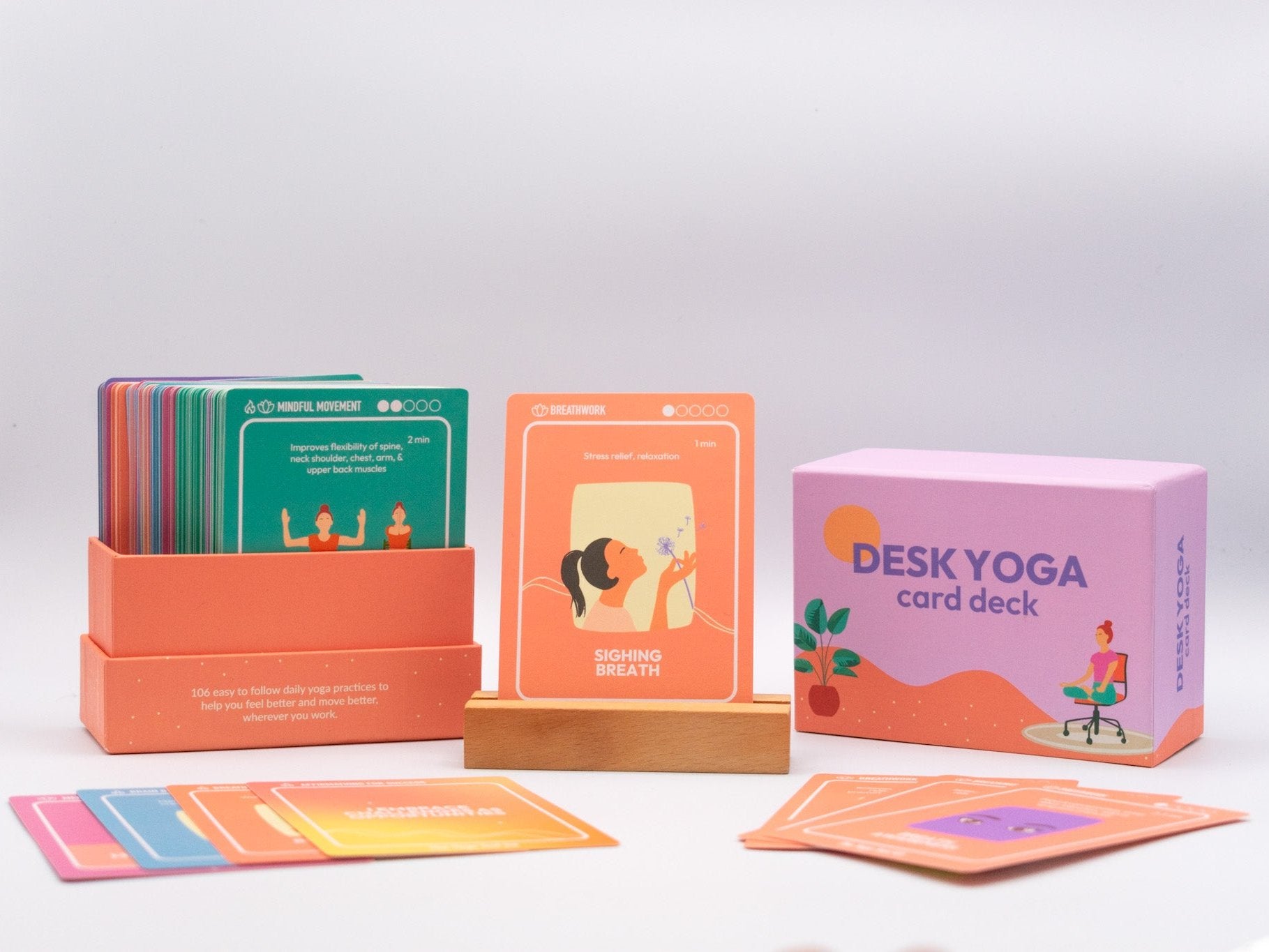 A box containing various desk yoga cards, with one yoga breathwork card "sighing breath" displayed on a wooden holder, alongside with the lid of the box removed and off to the side, showcasing the colorful deck, and a collection of colorful and diverse card designs from The Yoga Tool Kit. 