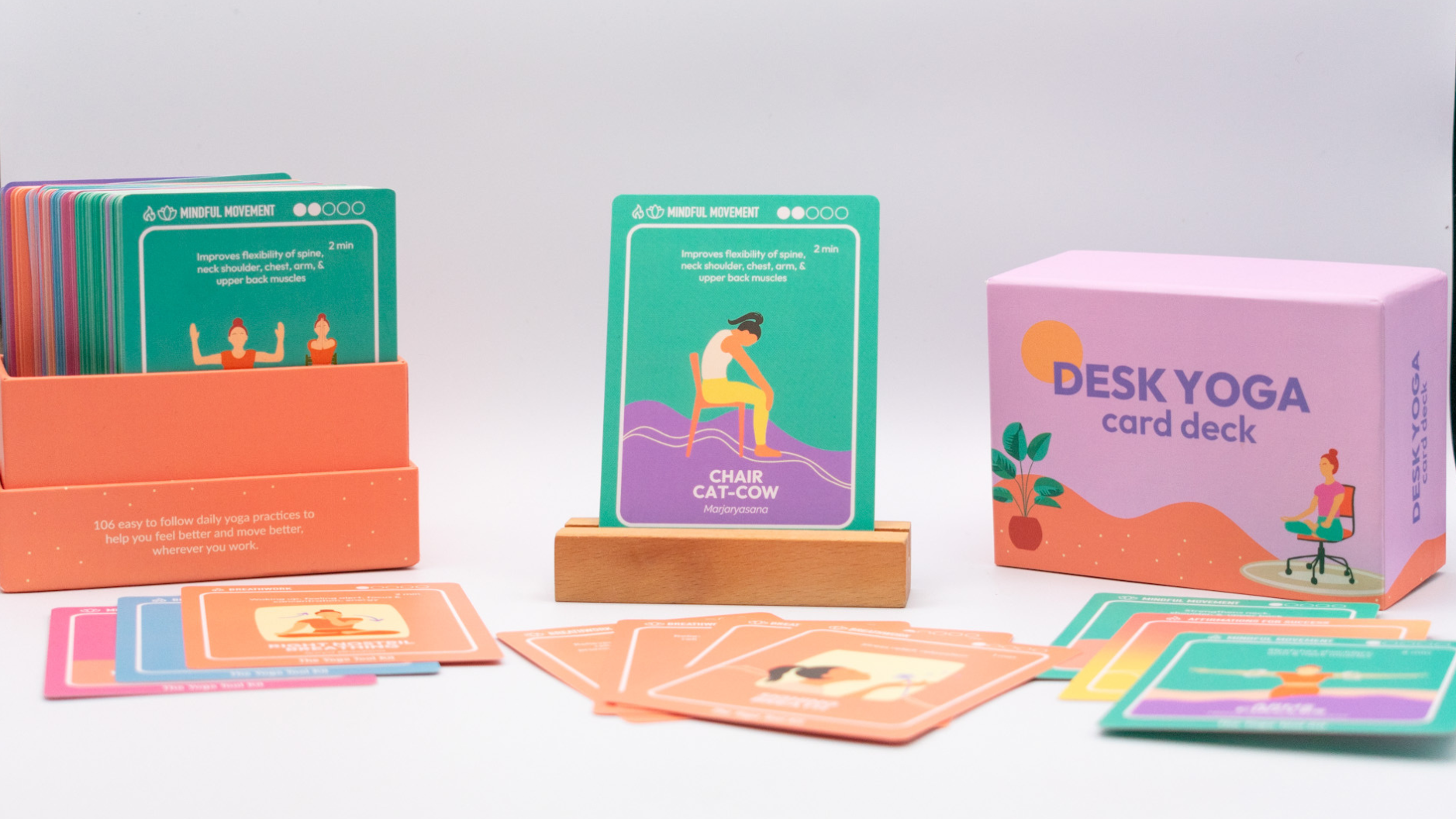 A box containing various desk yoga cards, with one yoga card "chair cat-cow" displayed on a wooden holder, alongside the lid of the box removed and off to the side, showcasing the colorful deck, and a collection of colorful and diverse card designs from The Yoga Tool Kit. 